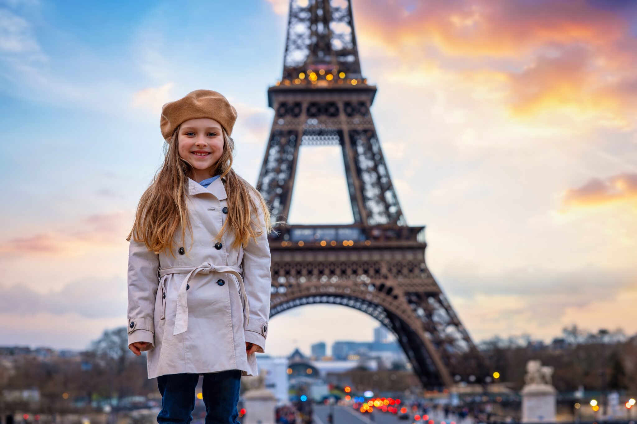 Portrait of a cute, blonde girl with a trench coat and a beret hat in front of the Eiffel Tower in Paris, France